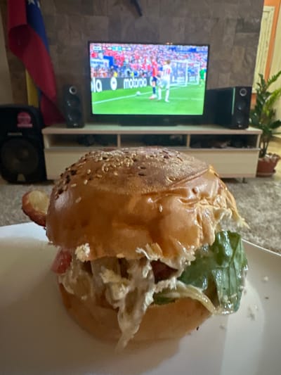 In a domestic setting, likely a living room in La Guardia, Venezuela, a close-up shot focuses on a hamburger placed on a white plate. The hamburger is substantial, with a sesame seed bun, visible lettuce, and what appears to be sauce or mayonnaise. In the background, a television displays a soccer match, with a crowd and players visible on the screen. To the left of the television, a flag, possibly the Venezuelan flag, is draped, featuring red, yellow, and blue colors with stars. Two black speakers are positioned on either side of the television, and a speaker unit is visible to the far left. A plant in a pot sits to the right of the television, and a piece of furniture, possibly a TV stand, is situated beneath the screen. The overall scene suggests a casual evening, possibly spent watching sports while enjoying a meal.