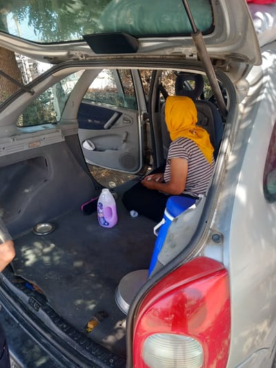 A person wearing a yellow headscarf and a striped shirt sits in the back of a silver hatchback car. The car is parked outdoors in what appears to be a rural or semi-rural environment, with trees and foliage visible in the background. The trunk of the car is open, revealing a blue cooler, a bottle of purple liquid, a metal dish, and other miscellaneous items. The car's interior is visible, including the rear seats and door panels. The scene is lit by natural daylight, casting shadows on the ground and the car. The location of Platani, Greece is not discernible from the image itself.