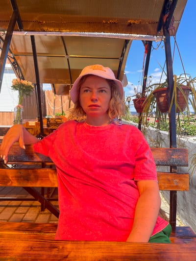 In the city of Belozersk, Russia, a blonde woman wearing a pink bucket hat and a bright pink t-shirt sits on a wooden bench under a translucent orange awning. The bench is weathered, with visible wood grain and some wear. She has her left arm resting on the back of the bench. Behind her, potted plants hang from a metal frame, and a glimpse of the sky and distant foliage can be seen. A vase of flowers sits on a table to her left. The ground beneath the bench appears to be paved with interlocking stones. The lighting suggests it is daytime, with sunlight casting shadows.