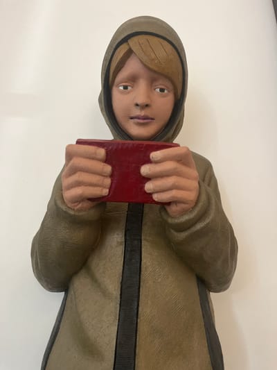 The image displays a sculpted figure, located in Galatina, Italy, depicting a youthful individual from the upper torso upwards. The subject possesses light brown hair visible beneath an olive-green hooded garment. The face exhibits a neutral expression, with painted eyes and lips. Both hands are sculpted holding a rectangular, deep red object horizontally at chest height. The olive-green garment features a distinct dark vertical stripe down its center. The figure is presented against an unadorned, light-colored wall, suggesting an indoor setting.
