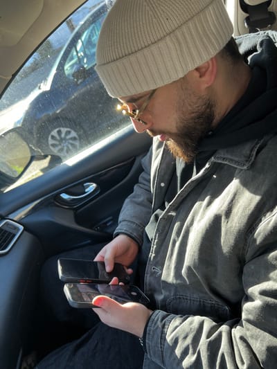 In Hérouville-Saint-Clair, France, a male individual is seated in the passenger seat of a vehicle, angled downwards. He wears a light beige knit beanie, gold-framed glasses, a dark hooded garment, and a faded dark grey jacket. Both hands hold black smartphones with dark screens. The top phone is held by his right hand; the bottom phone is held by his left hand, notable for its bright coral-painted thumb and index fingernails. Strong sunlight illuminates the scene, creating distinct highlights and shadows on the individual and the car's interior. Through the passenger window, a dark-colored vehicle is partially visible in the background.