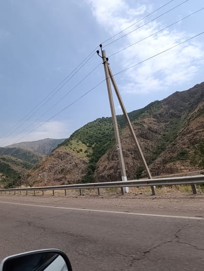 This is a photograph taken from a moving vehicle, likely on a highway, in a mountainous and somewhat arid region. The foreground shows a cracked asphalt road with a white line dividing lanes, and a section of a car's side mirror is visible in the bottom left corner. A metal guardrail runs along the side of the road, separating it from the rocky, vegetated mountains.

Dominating the middle ground and background are steep, rugged mountains. The slopes of these mountains are covered with sparse vegetation, predominantly green shrubs and trees, interspersed with exposed rock faces and dry, brownish earth. The sky is blue with scattered white clouds.

Several power lines stretch across the frame, supported by a prominent, angled concrete utility pole in the center-left of the image. Another, more distant power pylon can be seen on the far left side of the image. The overall impression is one of a scenic but somewhat remote, natural landscape with the presence of human infrastructure for power transmission.

The image's caption indicates the location as Saiyakka, Uzbekistan. This detail suggests the photograph captures a segment of the road network within or near this region of Uzbekistan, characterized by its mountainous terrain and infrastructure for electricity.