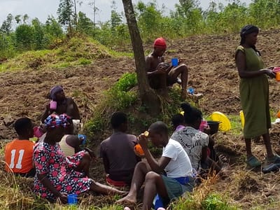 A group of people are gathered on a hillside that appears to be farmland. They are resting and eating or drinking from cups. Some are sitting on the ground, while others are standing. The group includes men, women, and children, some wearing colorful clothing, others more subdued tones. A tree provides shade for some, and farming tools and water containers are scattered around. The background consists of trees and vegetation.