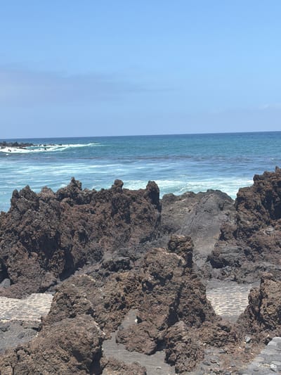 This is a vertical, eye-level, outdoor shot of a rocky coastline. The scene is dominated by dark, rough volcanic rocks on the right, with the ocean stretching out to the left. The water is a mix of turquoise and white, with visible waves and foam crashing against the shore. The sky is a clear, bright blue, suggesting it is a sunny day.
The rocks appear to be part of a natural cliff face or a man-made structure incorporating natural elements. There are areas of paving stones visible amongst the rocks, suggesting a pathway or viewing area. The texture of the volcanic rock is rough and porous. The ocean is active, with white caps on the waves, indicating a moderate surf.
The overall impression is one of natural beauty and ruggedness. The clear blue sky and the sparkling ocean suggest good weather, likely during the daytime. The location appears to be a coastal area, possibly a tourist spot given the paved sections. There are no people or man-made objects other than the paving stones. The image conveys a sense of a peaceful, yet wild, natural environment.
