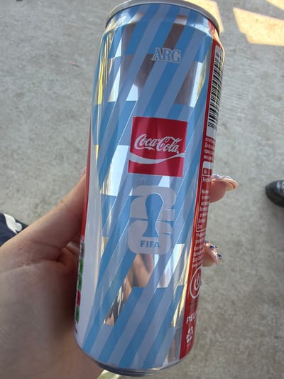 A hand with blue-jeweled fingernails holds a Coca-Cola can. The can is predominantly silver with light blue and white diagonal stripes. The Coca-Cola logo is in a red rectangle on the can, along with the word "FIFA." The can has text in a foreign language and a barcode. The background appears to be a light-colored concrete surface. The location is specified as Dinkata, Bulgaria.