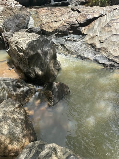 The image shows a close-up view of a rocky riverbed with murky, light green water. Large, weathered rocks of various sizes and textures dominate the foreground and middle ground. Some rocks are smooth and rounded, while others have rough, layered surfaces with visible striations. Sunlight casts dappled shadows on the rocks and water. In the background, a small cascade of water flows over some of the rocks. A few indistinct figures can be seen standing in the water near the cascade, suggesting this is a location people visit. The environment appears to be natural and undeveloped. This scene is located in the city of Fobur, Nigeria.
