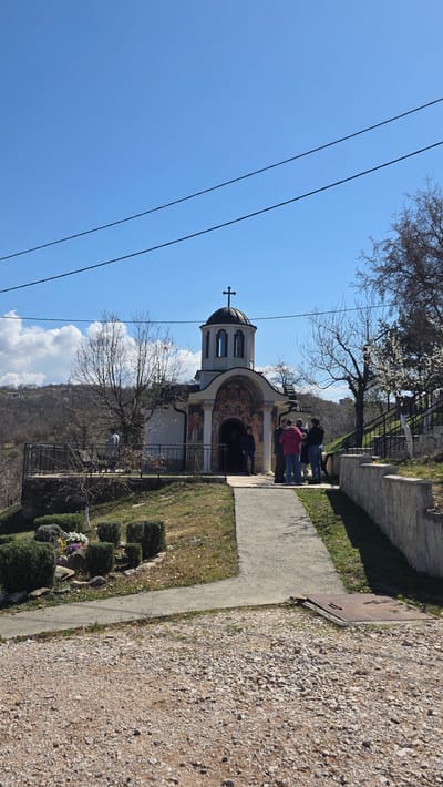 The image shows a small, ornate Eastern Orthodox church located in Elenovo, Bulgaria. A group of people is gathered at the entrance. The church has a dome topped with a cross, painted facades, and is set on a hillside. Power lines are visible against a clear blue sky with a few clouds. A paved path runs alongside the church, bordered by grass and small shrubs. A stone wall is further to the right of the path. The ground is covered with gravel.