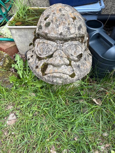 The image shows a weathered, stone ornament with decorative carvings, partially covered in moss, resting on the ground in Snailbeach, United Kingdom. The ornament is positioned next to a patch of green grass and dandelions. Adjacent to the ornament are gardening supplies, including a dark gray watering can, a blue tray, a gray container and a section of green garden hose. A weathered brick and moss are also visible in the frame.