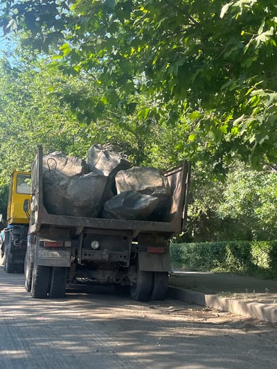 A truck is parked on a street, its bed filled with large rocks. The truck is yellow with a dark bed and visible tires. The rocks are a mix of grey and tan, with some appearing to be rough and uneven. The truck is positioned at an angle to the viewer, with the rear of the truck facing the right side of the frame. The scene is outdoors, with abundant green foliage from trees and bushes visible on the left and in the background. The sunlight suggests it is daytime, and the shadows indicate the sun is at a moderate angle, likely late morning or early afternoon. The presence of a sidewalk and paved road indicates an urban or semi-urban setting. The license plate on the truck reads "56 SU 706". The overall impression is a functional scene of a truck used for hauling heavy materials, set against a backdrop of natural greenery. There are no people visible in the frame. The weather appears to be clear and sunny.