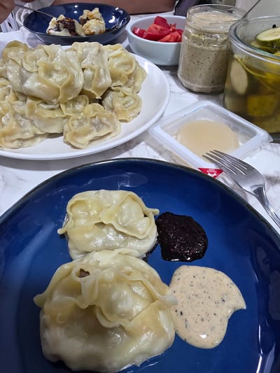 A close-up, overhead shot captures a meal on a marble-patterned surface. In the foreground, a vibrant blue plate holds two large, pale dumplings. A dollop of dark, jam-like condiment and a swirl of creamy, speckled sauce are served alongside the dumplings. A fork rests on the edge of the plate. Behind this, a white plate is piled high with more of the same dumplings. To the right, a small clear container holds a pale, gelatinous food item. Further back, a glass jar contains dill pickles, and another glass jar holds a creamy, speckled condiment. A small white bowl filled with chopped red tomatoes sits between the jars. The scene is dimly lit, suggesting an indoor setting, possibly a dining room. There are no discernible landmarks or elements that would indicate a specific location within Eumseong-eup, South Korea.