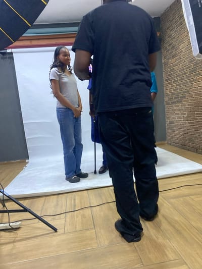 An indoor photography studio in Animashawun, Nigeria, is captured in this image, showing a photoshoot in progress or setup. In the foreground, a person stands with their back largely to the camera, wearing a black t-shirt and dark trousers, facing towards the right of the frame. They are positioned on a light-colored wooden floor.
Further back and to the left, a woman stands on a white seamless paper backdrop, which extends from the floor upwards. She is wearing a light grey t-shirt and blue jeans, with her hands clasped loosely in front of her. She has dark hair styled in braids and is looking towards the right, displaying a neutral to slightly pleasant expression. Her presence suggests she is the subject of the photoshoot.
Parts of other individuals are faintly visible in the deeper background, including someone wearing a blue top. A thin black electrical cord stretches across the wooden floor. On the far left, the frame of a large softbox or similar lighting equipment is visible. Another light stand is partially visible on the right. A brick-patterned wall is seen above the white backdrop, creating an industrial accent in the studio space. The word "Canon" is faintly visible on a strap or bag near the feet of the person in black.
