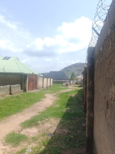 The image depicts a narrow dirt road in Ibo, Nigeria, flanked by concrete walls on both sides. The wall to the right is taller and has barbed wire on top. The road is grassy in parts, with evidence of wear from use. In the distance, buildings with various roof styles are visible, including one with a green roof and solar panels. A small hill is seen on the horizon. Two individuals are walking down the road in the distance. The sky is partially cloudy.