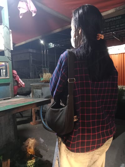 A person wearing a red and black plaid shirt, a white face mask, and light-colored pants stands facing away from the camera. They have a dark-colored shoulder bag with a strap that crosses their chest. The person is standing on a paved surface, possibly a street or sidewalk, at night. In the background, there are food stalls or carts, with skewers of food visible on one. A sign overhead reads "KOPERASI MAH KALIWUN". The lighting suggests it is nighttime, possibly a busy street food area. The overall scene is dimly lit, with the focus on the person and the surrounding environment.