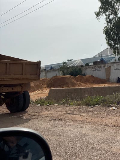 In the city of Ban, Nigeria, this image depicts a construction site. A large, weathered dump truck is parked on the left side, its bed filled with sand. In the background, a low concrete wall runs horizontally, topped with barbed wire. Behind the wall are several industrial-looking buildings with corrugated metal roofs, one of which appears to be a warehouse or factory. Piles of sand are scattered around the site, suggesting ongoing construction or material storage. A person is visible sitting near the right edge of the wall, partially obscured. The ground is a mixture of gravel and dirt, with sparse vegetation and some litter present. The sky is overcast, contributing to the muted lighting of the scene.