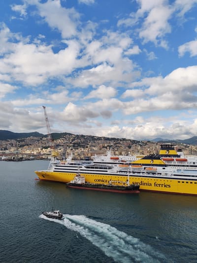 The image shows a port in Genoa, Italy. In the foreground, a small white pilot boat is moving through the water, leaving a trail of white wake. Behind it are two large yellow and white ferries, labeled "Corsica Ferries" and "Sardinia Ferries". One ship is docked, while the other is in the process of docking. A small black oil tanker is docked beside the large yellow and white ferry. In the background, a hillside city is visible, with buildings and a crane dotting the landscape under a sky filled with white clouds.