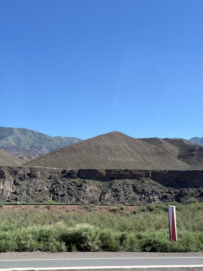 This media file depicts a vast mountainous landscape under a clear, bright blue sky, suggesting a sunny day. The scene is oriented vertically, with the sky occupying the left portion and the earth the right. The terrain is characterized by steep, rugged mountains with textured slopes covered in sparse vegetation and rocky outcrops. The foreground on the right features a paved road with white markings, bordering a strip of lush green grass and some scattered bushes. A red and white marker is visible on a pole along the edge of the road, likely a directional or safety sign. The overall impression is one of a remote, natural environment, possibly a roadside view in a mountainous region. No people or specific activities are depicted. The lighting and clear sky suggest it is daytime, likely mid-morning or afternoon. The image captures a sense of open space and natural grandeur.