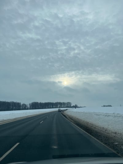 The image depicts a winter road scene, likely on a highway or major thoroughfare in or near Shelepino, Russia. The sky is largely overcast with light grey stratocumulus clouds, through which a diffused, yellow sun is visible in the upper-middle right quadrant. The road, observed from a low-angle perspective suggesting an interior vehicle viewpoint, is a two-lane asphalt surface, appearing clear of snow but potentially wet. White lane markings are visible. Snow banks, discolored with brown and black road grime, line both shoulders of the road. Beyond these banks, the surrounding landscape is uniformly covered in pristine white snow. A dense line of leafless deciduous trees forms a dark treeline along the left side of the road, extending into the distance. On the right, snow-covered fields recede, with sparse clusters of bare trees visible. Several vehicles are observed traveling in the same direction as the vantage point, including a white van in the mid-distance and at least one other passenger vehicle further ahead. No individuals are explicitly discernible outside of vehicles. The overall environment suggests a cold, winter day in a rural or exurban setting.
