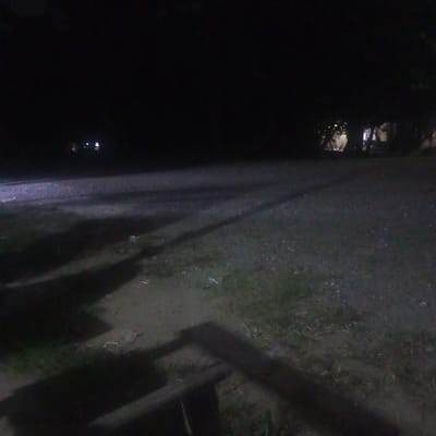A nighttime scene in what appears to be a park or open area. The ground is a mix of dirt, gravel, and sparse grass. The bottom of the frame is filled with the shadows of wooden beams, suggesting a bench. In the background, faint lights indicate structures or buildings. In the distance, there are sparse lights, likely from houses or street lamps. Based on the location provided, it is likely a park or recreational area in Matangmeunye, Indonesia. No people are visible in the image.