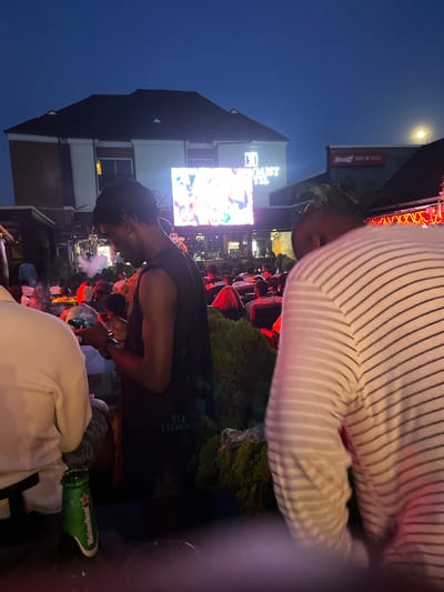The image depicts an outdoor gathering in Offatedo, Nigeria. People are seated and standing in what appears to be a restaurant or bar setting. A large screen displays a broadcast, possibly a sporting event. Foregrounded are individuals, one in a striped shirt and another in a black tank top, who are partially obstructing the view of the scene. A green Heineken beer bottle is visible in the lower right corner of the frame. Streetlights illuminate the area. Buildings, including a "MIST" hotel and a Budweiser advertisement are visible in the background.