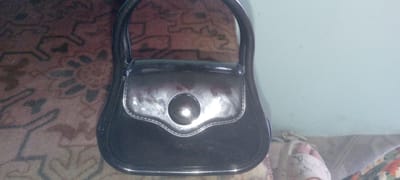 A black patent leather handbag with a rounded shape and a decorative button closure is positioned on a patterned rug. The handbag has a single, curved handle. The rug appears to be in a dimly lit room, with a pale blue wall visible in the background. The image is taken from a slightly elevated angle, looking down at the handbag. The location is specified as Marhamat, Uzbekistan.