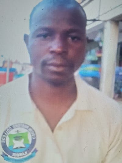 The media file is a close-up, rotated portrait of a young person. The subject is looking towards the camera with a neutral expression. They are wearing a light-colored uniform with a circular emblem on the left chest. The emblem features the letters "KYBMS COLLEGE" and a green symbol in the center. The background is blurred and appears to be indoors, possibly a classroom or office, with hints of colorful objects. The lighting suggests it is daytime. The image quality is low, with visible pixels and some artifacts. The overall impression is a casual, possibly candid, photograph.