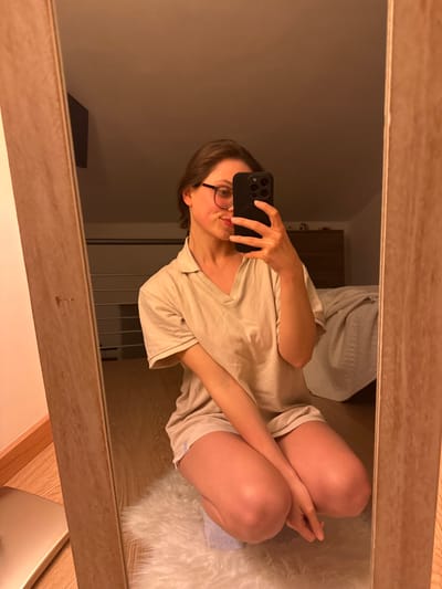 An image captured via a mirrored reflection depicts a female individual in a residential interior located in Cava de' Tirreni, Italy. The subject, a woman with brown hair and dark-rimmed glasses, is shown from the chest down, crouching on a white, shaggy rug. She is wearing a light beige short-sleeved, collared V-neck top and matching shorts. Her right hand holds a dark-colored smartphone, equipped with a pop socket or similar grip accessory, angled to photograph her reflection in the full-length mirror. The mirror has a light-colored wooden frame and is positioned on a wooden floor. In the background, reflected within the mirror, elements of the room are visible: a light-colored wall, a horizontal metallic railing or shelving unit, a chest of drawers or dresser with dark handles, and a portion of a bed with light-colored bedding. Artificial warm lighting illuminates the scene.