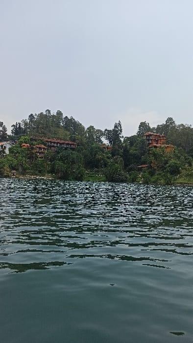 The scene, located in Bwishyura, Rwanda, presents a body of water in the foreground under an overcast sky. The lower two-thirds of the image depict dark greenish-blue water with a rippled surface, exhibiting patterns of light and shadow. Above the waterline, a verdant hillside rises, densely covered with various green trees and thick foliage. Multiple buildings are situated on this hillside, integrated within the natural vegetation. On the left side, a cluster of multi-story structures with white facades and reddish-brown tiled roofs is visible. Towards the right, additional buildings, also featuring reddish-brown tiled roofs and natural-toned walls, are nestled among the dense trees. The upper third of the image is occupied by a uniform, light gray sky. No human figures or discernible actions are present.