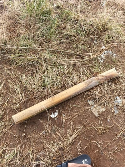A hollow bamboo section rests on a mixed terrain of dry grass, green patches, and bare earth. Scattered trash, including small plastic pieces, lies around. Thin twigs and branches crisscross the scene. The image is shot at ground level, capturing the rough texture of the natural elements and emphasizing the discarded bamboo's linear form. A partial view of a person's footwear is at the bottom.