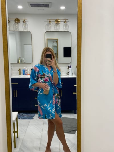 A full-length mirror selfie captures a female subject standing barefoot in a residential bathroom. The subject partially obscures her face with a light-colored smartphone held in her right hand, positioned for a reflection capture. She wears a mid-thigh length, light blue satin-like robe patterned with pink flowers and green/purple peacock plumage. Her left hand holds a clear cup containing a light brown beverage with a white layer on top. A blue and silver beaded bracelet is visible on her right wrist. The bathroom features dual white rectangular sinks with gold-colored faucets and dark blue vanity cabinets with gold-colored bar handles, situated under white countertops. Above each sink, a gold-toned light fixture with three clear glass shades is mounted. Various toiletries are present on the countertops. The floor is composed of white square tiles with gray veining. A blue and white striped mat lies to the left, partially beneath a white stool with gold legs. A gray bathmat is positioned on the floor to the right. The walls and ceiling are white, with a rectangular ventilation grate and a recessed light visible on the ceiling. The specific city of Ridgefield, United States, is not discernible from the image; however, the setting is consistent with a private residence.