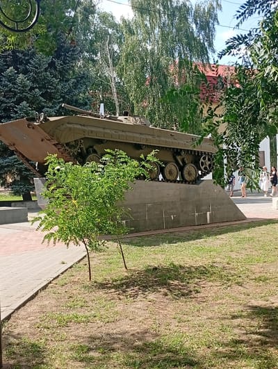 An olive green armored personnel carrier monument is displayed on a concrete pedestal in a park in Pokrov, Ukraine. The vehicle is positioned at an upward angle, facing left, with its tracks and side visible. The pedestal is inscribed with text, though it is not clearly legible in the image. Two small green trees are situated in front of the pedestal. In the background, lush green trees and the red roof of a building are visible under a bright sky. Several people, including women and a child, are walking on a paved walkway to the right of the monument. The ground in the foreground is covered with grass and dirt.