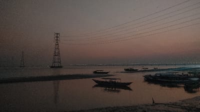 The image captures a wide-angle view of a body of water at twilight in Kumarvog, Bangladesh. The sky transitions from a dark gray at the top to a muted pinkish-orange glow near the horizon. Multiple parallel power transmission lines extend horizontally across the upper mid-ground. Two lattice-steel transmission towers are visible rising from the water; one is prominent in the mid-left, supporting the power lines, and a smaller, fainter tower is located further left in the background. The water's surface reflects the sky's colors, appearing dark with horizontal strips of shallower water or sand. Several boats are present: a long, narrow boat with a canopy is positioned in the central mid-ground, and a cluster of similar boats are gathered towards the right foreground, near a dark, uneven bank. In the far distance, a bridge spans the water, illuminated by a series of evenly spaced lights. No discernible human figures or specific actions are visible on the boats or shoreline.