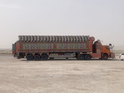 A large, brightly decorated truck is parked on a flat, dusty landscape under a hazy sky. The truck is loaded with many cylindrical objects wrapped in gray material. Ornate floral and geometric designs cover the truck bed and cab. A white car is parked near the truck's front. The overall scene suggests a transport vehicle in a dry, possibly arid environment.