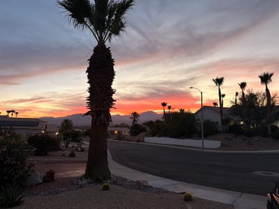 The image depicts a residential street in Desert Hot Springs, California, during a vibrant sunset. In the foreground, a large palm tree trunk dominates the left side of the frame, with its fronds silhouetted against the colorful sky. The ground is covered in gravel, interspersed with decorative rocks and a few barrel cacti. A paved road curves through the scene, lined by a sidewalk and landscaping that includes more gravel, rocks, and low-lying plants.
To the left of the road, a house with solar panels on its roof is visible, surrounded by vegetation and some terracotta pots. Further down the street, several other houses can be seen nestled amongst trees and landscaping. A street lamp stands prominently on the right side of the road, its light faintly illuminating the scene.
In the background, a range of mountains forms the horizon, bathed in the warm hues of the setting sun, which paints the sky in shades of orange, pink, and purple. Other palm trees are scattered throughout the scene, some silhouetted against the sky. The overall atmosphere is serene and depicts a typical desert suburban landscape at dusk. There are no people visible in the image.