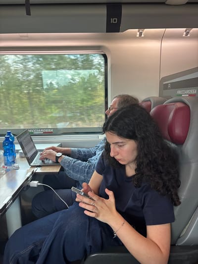 The scene depicts the interior of a Frecciarossa high-speed train carriage, likely traveling through or near Castelfranco Emilia, Italy. Two individuals are seated. In the foreground, a young woman with long, dark, curly hair, wearing a dark blue t-shirt and dark blue pants, is engrossed in a silver smartphone held in both hands. She has rings on her left hand and a silver bracelet on her right wrist. Behind her and to the left, an older man with short, gray hair and eyeglasses, dressed in a blue plaid shirt, is seated at a wooden fold-out table next to a window. He is actively typing on an open silver laptop. A blue plastic water bottle, a clear glass, and a small metallic container rest on the table. A white charging adapter with a cable is plugged into a power outlet below the table. The man wears a watch on his left wrist. Through the train window, a blurred exterior shows green trees, consistent with a train in motion through a natural landscape. The train interior features grey seats with red headrests and an overhead panel marked with the number "10".