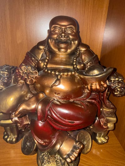 A golden-bronze statue of a laughing Buddha sits on a wooden shelf. The Buddha is depicted as plump with a smiling face, wearing a beaded necklace and a red garment. He is seated on a platform decorated with what appear to be Chinese coins. To his left, he holds a bulging sack, and to his right, he holds a gold ingot. The background is a warm brown wooden surface, likely part of a shelf or furniture. There is a light source from the right, casting a soft glow on the statue and creating a shadow. The statue is centrally framed in the image. The location is not identifiable from the image.