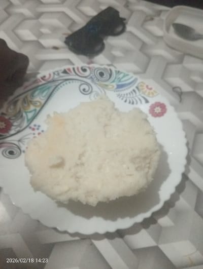 The scene, located in Kapkures ward, Kenya, depicts an indoor setting, likely a table. A white, scalloped-edge plate with colorful decorative patterns, including swirls and floral designs, is centrally positioned. On the plate rests a mound of white, granular, cooked food, possibly rice or a similar starchy staple. To the upper left of the plate, a pair of dark-framed sunglasses with blue-tinted lenses lies on the surface. Partially visible in the upper right corner is a portion of a white, possibly ceramic, bowl or container. A dark-skinned hand or arm is partially visible on the far left. The surface beneath all objects is light-colored with a repeating three-dimensional geometric pattern, possibly a tiled countertop or tablecloth. A digital timestamp "2026/02/18 14:23" is visible in the lower left corner of the image.