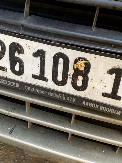 A close-up, slightly angled, eye-level outdoor shot shows a dirty license plate mounted on a vehicle's grille. The license plate is white with black numbers and text. The visible portion of the license plate reads "26108 1" followed by smaller text that includes "Verkauf", "Castroper Hellweg 372", and "44805 BOCHUM". A dead insect is positioned on the number "8". The grille is black plastic and appears dusty. The lower part of the image shows a silver-colored bumper with horizontal slats, also dusty and with some debris. The background is dark and out of focus. There is no visible indication that this scene is located in Baraki, Algeria.
