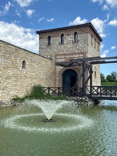 The image depicts a historical structure, likely a fortress or castle, in Neofit Rilski, Bulgaria. The building is constructed of stone and brick, with arched windows and a large arched entrance. A wooden drawbridge extends from the structure over a body of water, where a fountain is actively spraying water. The water is green, suggesting it might be a pond or a lake. The sky is blue with scattered white clouds. There is a wooden fence along the edge of the water. The overall scene evokes a sense of history and tranquility, with the natural elements of water and sky complementing the ancient architecture. No people are visible in the image.