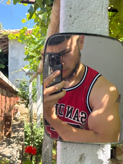 A young man with dark hair and a beard takes a selfie in a square mirror, reflecting his upper body. He is wearing a red basketball jersey with white trim. His right arm is visible, and his left arm holds a black smartphone, pointed towards the mirror. The reflection shows a background of lush green foliage, a brick wall on the left, and a light-colored building with a red-tiled roof on the right. A bright red flower is visible near the bottom of the reflection. The overall scene suggests an outdoor setting, likely a residential area in Konop, Bulgaria, given the architectural style of the buildings and the surrounding vegetation. The lighting indicates a sunny day.