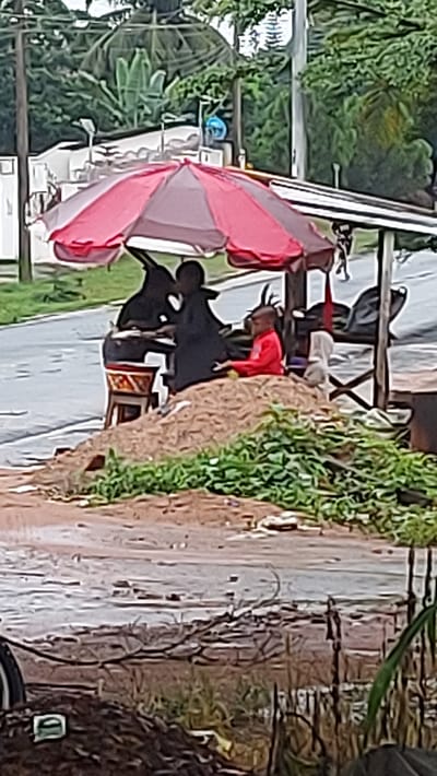 In Igbere, Nigeria, a boy in a red shirt sits on a pile of dirt under a red and gray umbrella. Beside him, another person, dressed in black, sits behind a table. A second umbrella, also red and gray, is positioned to the left, partially obscuring the figures and table. In the background, lush green vegetation and trees dominate the scene, with a paved road visible to the left. The ground in the foreground is wet and muddy, with some debris scattered about, suggesting recent rain.