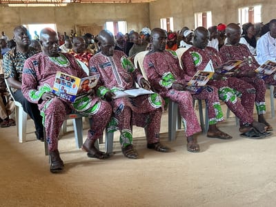 The image shows an indoor gathering, likely a church service or a meeting, in Kufana, Nigeria. Several men, predominantly older, are seated in plastic chairs, facing forward. They are dressed in matching maroon and white patterned traditional attire. Many are holding and reading booklets or programs. The room appears to be sparsely furnished with a dirt floor and basic walls. Natural light enters through windows. The overall atmosphere suggests a formal or religious occasion.