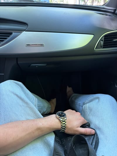 The image shows a person's legs and arm inside a car. The person is wearing light blue jeans and a watch with a metal band and a black face. Their hand is resting on a black bag with a textured pattern. The car's interior is visible, including a silver trim piece on the dashboard with the word "quattro" embossed on it, and an air vent. The scene appears to be set during the day, with natural light filtering into the car. The overall mood is casual. There is no clear indication of the weather or specific emotions. The location context is Orhei, Moldova.