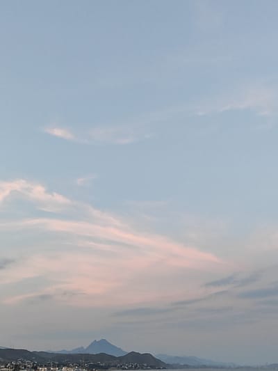 This media file depicts a scenic view of El Campello, Spain, likely captured during the late afternoon or early evening as indicated by the soft, pastel colors in the sky. The sky is a dominant feature, displaying hues of pale blue with streaks of pink and orange from the setting sun, suggesting a peaceful and serene atmosphere. wispy clouds are scattered across the upper portion of the frame.
Below the sky, a silhouette of hills and mountains forms the horizon line. These are not sharply defined, hinting at a distance. Nestled at the base of these hills, a town or village is visible, with lights twinkling, suggesting it is either dusk or the artificial lights are beginning to illuminate. In the foreground, the ocean or a large body of water is present, though the water itself is not clearly detailed. Some boats, possibly yachts or sailboats, are docked or anchored near the shore, identifiable by their masts. The overall setting appears to be coastal, with the town built along the shoreline and extending up the gentle slopes of the hills.
There is no visible text in the image. The scene conveys a sense of tranquility and natural beauty, typical of a coastal town at the end of the day. The absence of people or specific activities makes it a general landscape shot, focusing on the interplay of light, sky, land, and water.