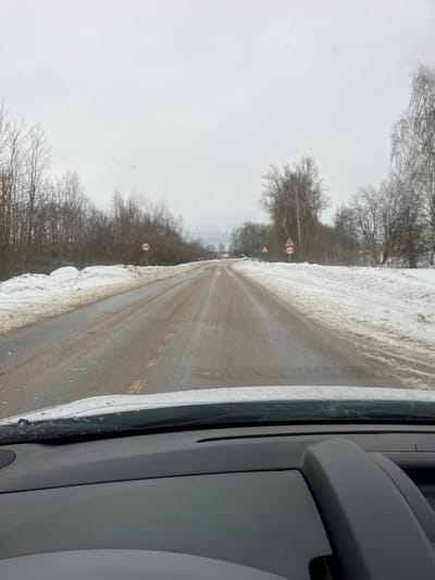 The image depicts a two-lane asphalt road in Rogachovo, Russia, during winter, viewed from the interior of a vehicle looking forward through the windshield. The immediate foreground shows the vehicle's dashboard and the base of the windshield, partially covered with ice or melted snow. The sky is overcast and uniformly gray. The road surface is wet, appears slushy or partially frozen, and is marked with streaks of dirt or sediment. Faint yellow dashed lines are visible in the center of the road, indicating two-way traffic. Snowbanks, discolored with dirt, line both sides of the road. On the left, a dense line of bare deciduous trees and some evergreens parallels the road. A red and white circular prohibitory road sign, resembling "No Entry for Vehicles with Trailer," is mounted on a pole on the left roadside. Further down the road on the right, multiple triangular warning signs with red borders are visible, one depicting a winding road and another possibly indicating a rough road or other hazard. Another circular prohibitory sign is also present on the right. A light-colored vehicle, possibly white, is faintly visible in the distance on the road, moving away from the viewer. Power lines are discernable in the middle distance on the left side, above the trees. No individuals are visible.