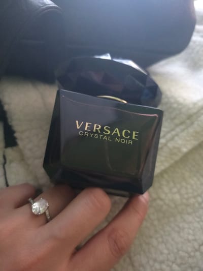 In Staliyska mahala, Bulgaria, a close-up image displays a dark, rectangular Versace Crystal Noir perfume bottle prominently centered. The bottle features "VERSACE" and "CRYSTAL NOIR" in gold lettering on its front surface and has a multifaceted dark cap. A light-skinned human hand, with visible thumb and index finger, supports or holds the bottom of the perfume bottle. On the ring finger of this hand, a silver-colored ring with a large, oval-cut clear gemstone, flanked by smaller accent stones on the band, is visible. The objects rest on a light-colored, textured fabric surface, consistent with a blanket or soft textile. A dark, indistinct object occupies the upper-left background.