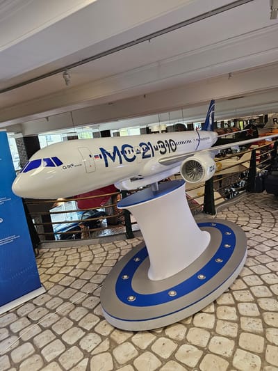 The image shows a model of an MC-21-310 aircraft in what appears to be a museum or exhibition hall. The aircraft is painted white with blue accents and markings. The aircraft model is suspended above a white stand with a circular grey and blue base. The floor is paved with light-colored stones. In the background, there is metal railing and other museum exhibits are visible. The location is likely in or near Архангельское, Russia, given the Cyrillic markings on the aircraft.