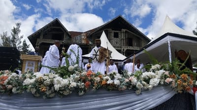 The image shows a group of people, primarily men, dressed in white robes, standing behind a white table adorned with a large floral arrangement. Several of the men hold various objects, including a shield, spears, and a cross. The event is being held outdoors, with a large white tent, a stone building with a wooden roof, and lush greenery visible in the background. The sky is partly cloudy and blue. The location is Rukoko, Democratic Republic of the Congo, at coordinates -1.624, 29.258. The scene is reminiscent of a religious ceremony. The men in the foreground appear to be participating in a ritual or procession, while others are watching and engaging in conversations.