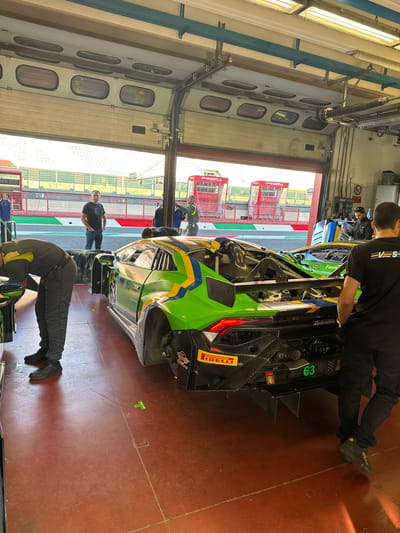 The image depicts a garage area at the Mugello Circuit in Scarperia, Italy, likely during a motorsport event. In the foreground, a green and yellow race car, possibly a Lamborghini, is parked. A person in a grey racing suit is bent over the car, appearing to work on it. Another person in a black t-shirt and dark pants stands to the right, facing away from the camera and looking at the race car. Further back, two other individuals are visible near the open garage doors. The garage doors themselves are elevated, revealing a track environment with tiered seating and red structures beyond. The floor of the garage is a reddish-brown color, and the walls are a light grey. There are various tools and equipment visible within the garage space. The overall scene suggests a pit stop or maintenance activity for the race car.