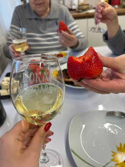 The image shows a person holding a strawberry with a bite taken out of it, poised above a glass of white wine. Another person in the background is holding a similar strawberry and a wine glass. A white plate with a sunflower design and a fork is visible on the right side of the image. The scene appears to be indoors, possibly during a meal or social gathering. The location is not discernible from the image. The city of Bashur, Russia cannot be confirmed.