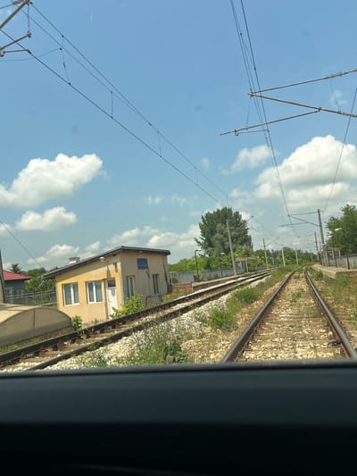 The image displays a sunlit railway scene, captured with a significant tilt as if rotated clockwise. It appears to be taken from inside a train or tram, looking out a window, with a dark vertical frame visible on the far right. Multiple railway tracks, covered in gravel ballast with some green vegetation and sparse red flowers, run diagonally from the foreground towards the distance.

Above the tracks, against a bright blue sky, an extensive network of overhead electrical lines and support structures is visible. These catenary systems feature thick wires, insulators, and metal support bars. Scattered white cumulus clouds punctuate the blue sky, indicating clear and sunny weather, likely during the daytime.

To the right of the tracks, a two-story, light yellow building with multiple windows and a white door stands. Some windows are fitted with white Venetian blinds. A small, rectangular blue sign with illegible text is mounted on the upper facade of this building. Beyond the building and along the railway line, dense green trees and foliage are visible. Further down the tracks, utility poles support the overhead lines. The scene depicts a typical railway environment, possibly near a station or siding in Madara, Bulgaria.